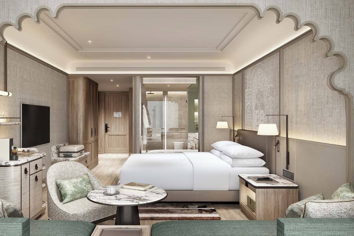 Une chambre d’hôtel moderne dotée d’un grand lit, d’un coin salon avec une table ronde, d’une télévision et d’une salle de bain visible à travers une porte en verre.