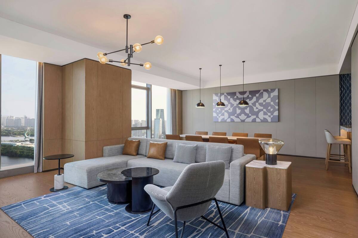 Ein modernes Wohnzimmer mit einem Ecksofa, einem runden Couchtisch und einem gemusterten Teppich. Über der Sitzecke hängen Pendelleuchten und große Fenster bieten Blick auf die Stadt.
