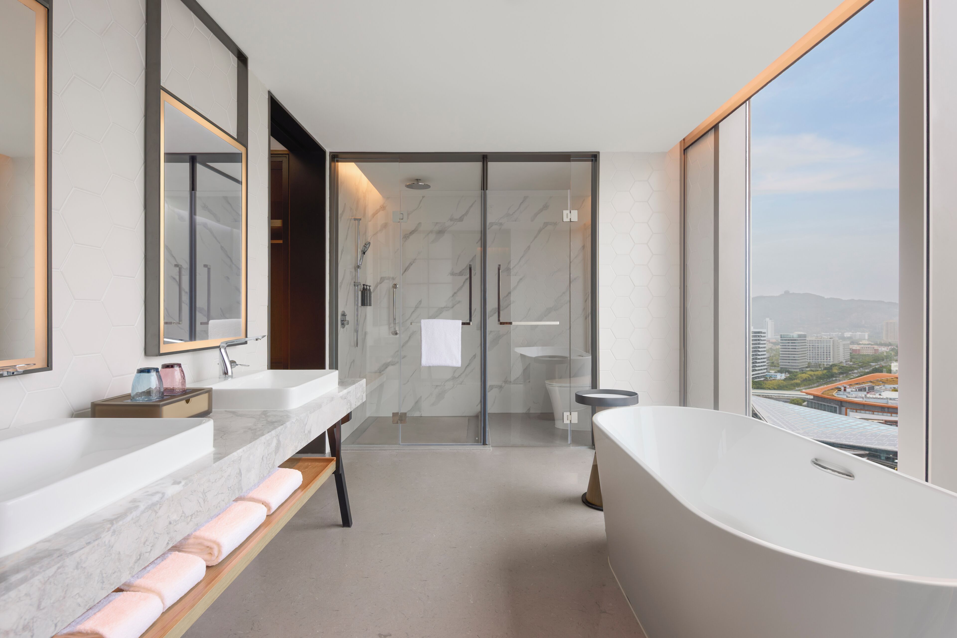 A modern bathroom with a large bathtub, a glass shower enclosure, and a city view.