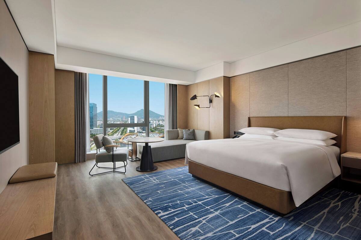 Ein modernes Hotelzimmer mit einem großen Bett, einer Sitzecke mit Tisch und Blick auf die Stadt durch große Fenster.