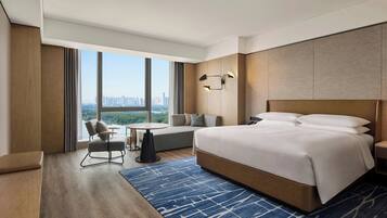 A modern hotel room with a large bed, a seating area with a sofa and a small table, and a view of the cityscape through large windows.
