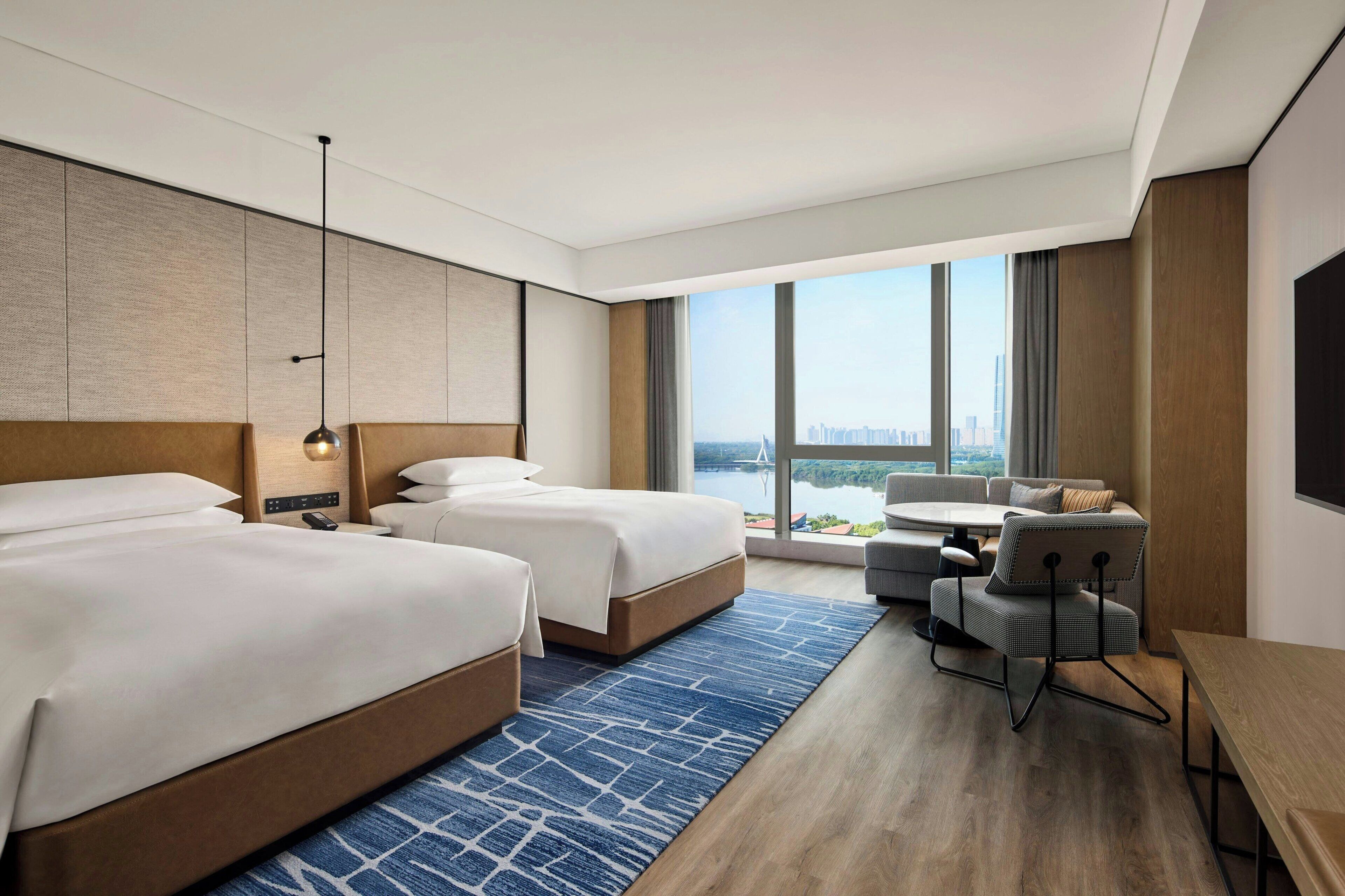 A hotel room with two beds, a sofa, a dining table, and a large window offering a city view.