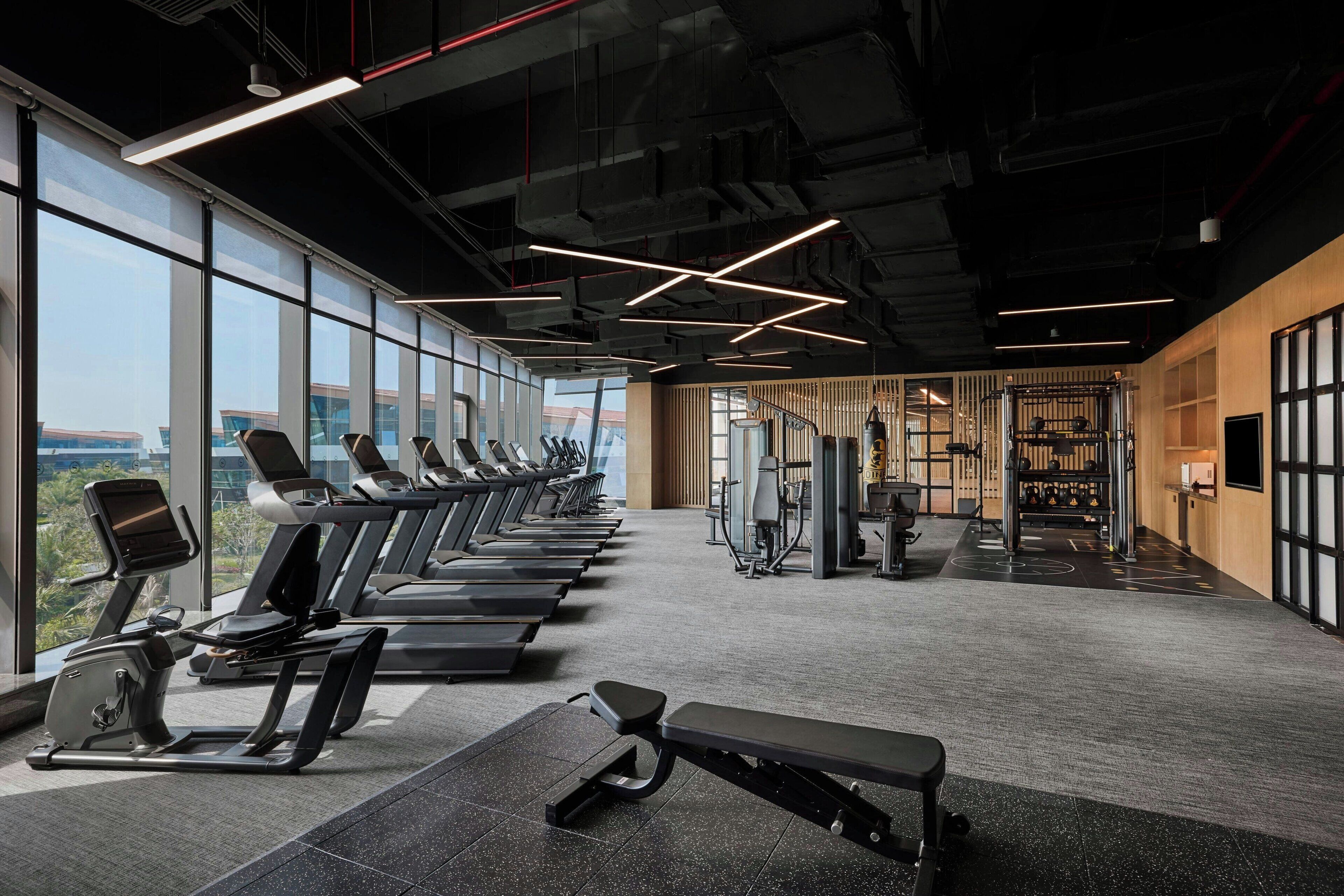 Une salle de sport équipée de tapis de course, de bancs d’exercices et d’appareils de musculation, avec de grandes fenêtres et une vue sur la ville.