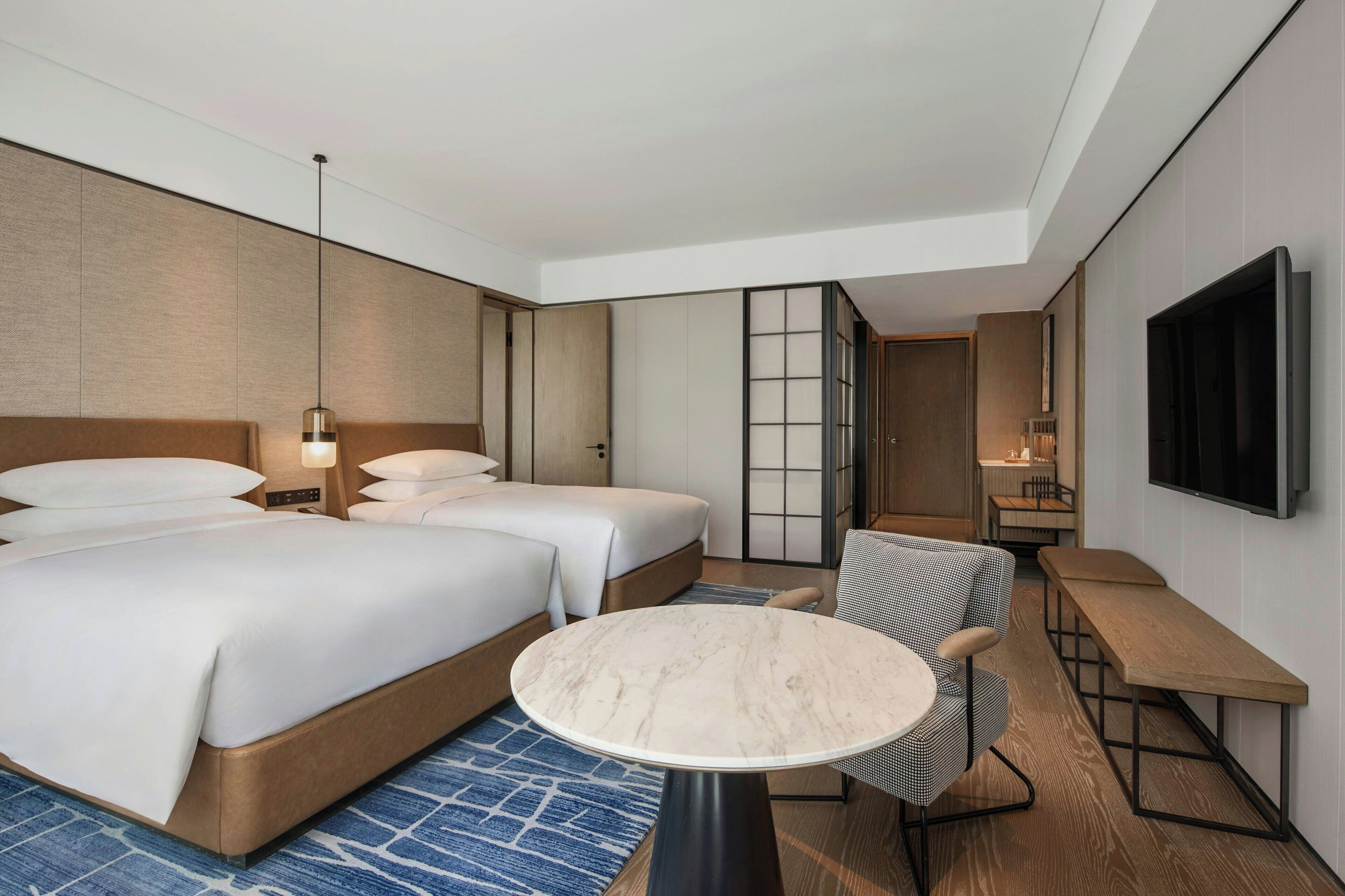 A hotel room with a large bed, two bedside tables, a round table, a chair, a TV, and a sliding door leading to a bathroom.