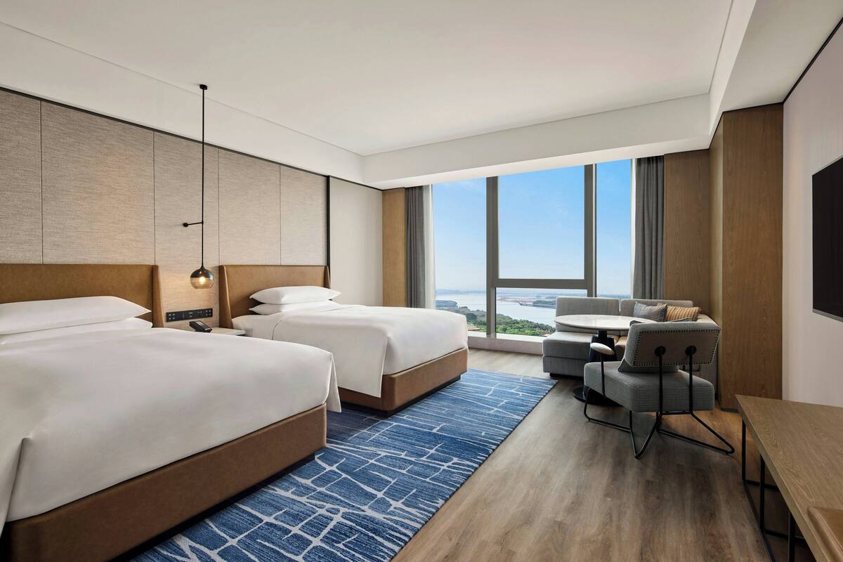 Ein Hotelzimmer mit zwei Betten, einem Schreibtisch, einem Stuhl und einem großen Fenster mit Blick auf die Stadt.