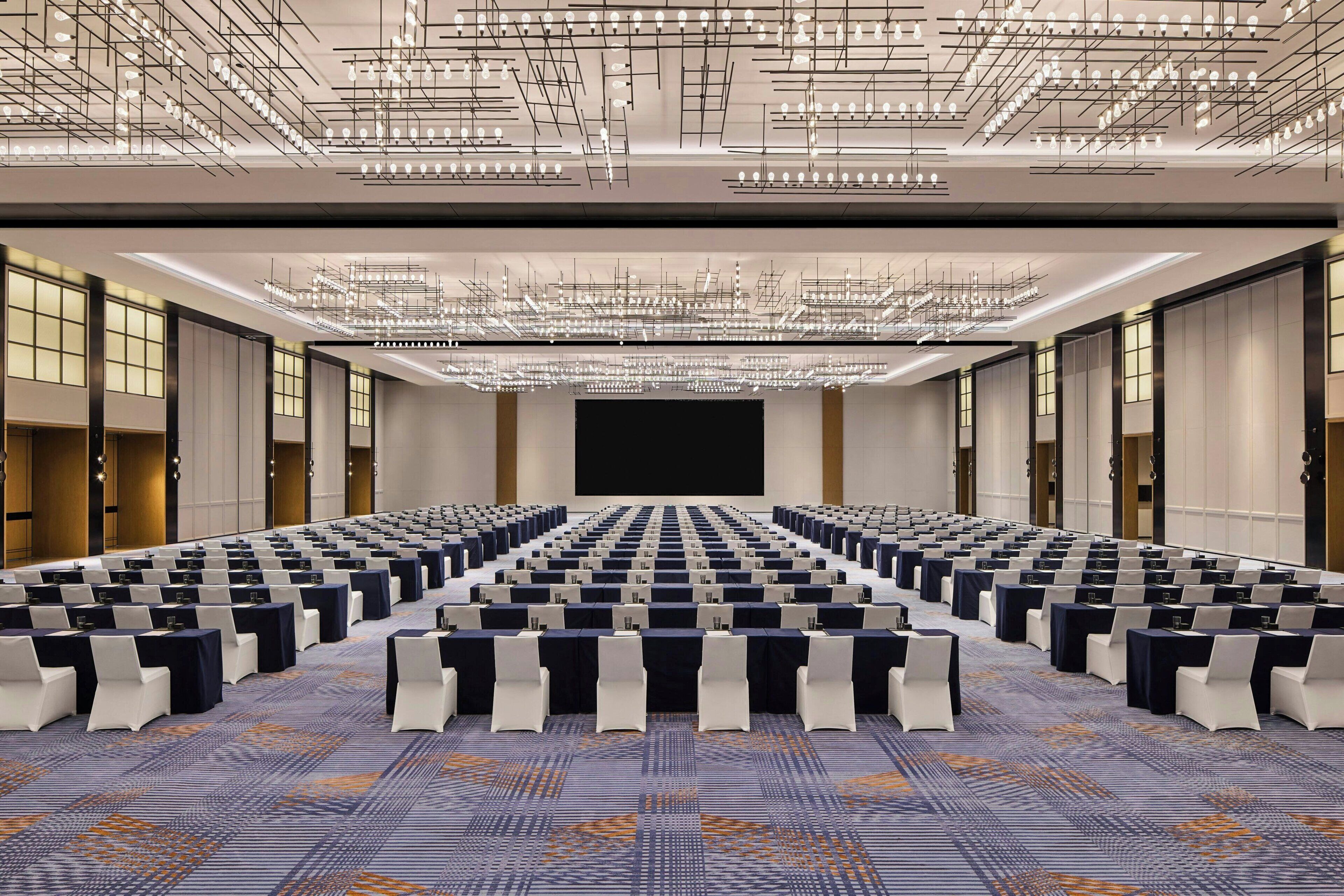 A large conference room with rows of tables and chairs, a large screen at the front, and a complex ceiling light fixture.