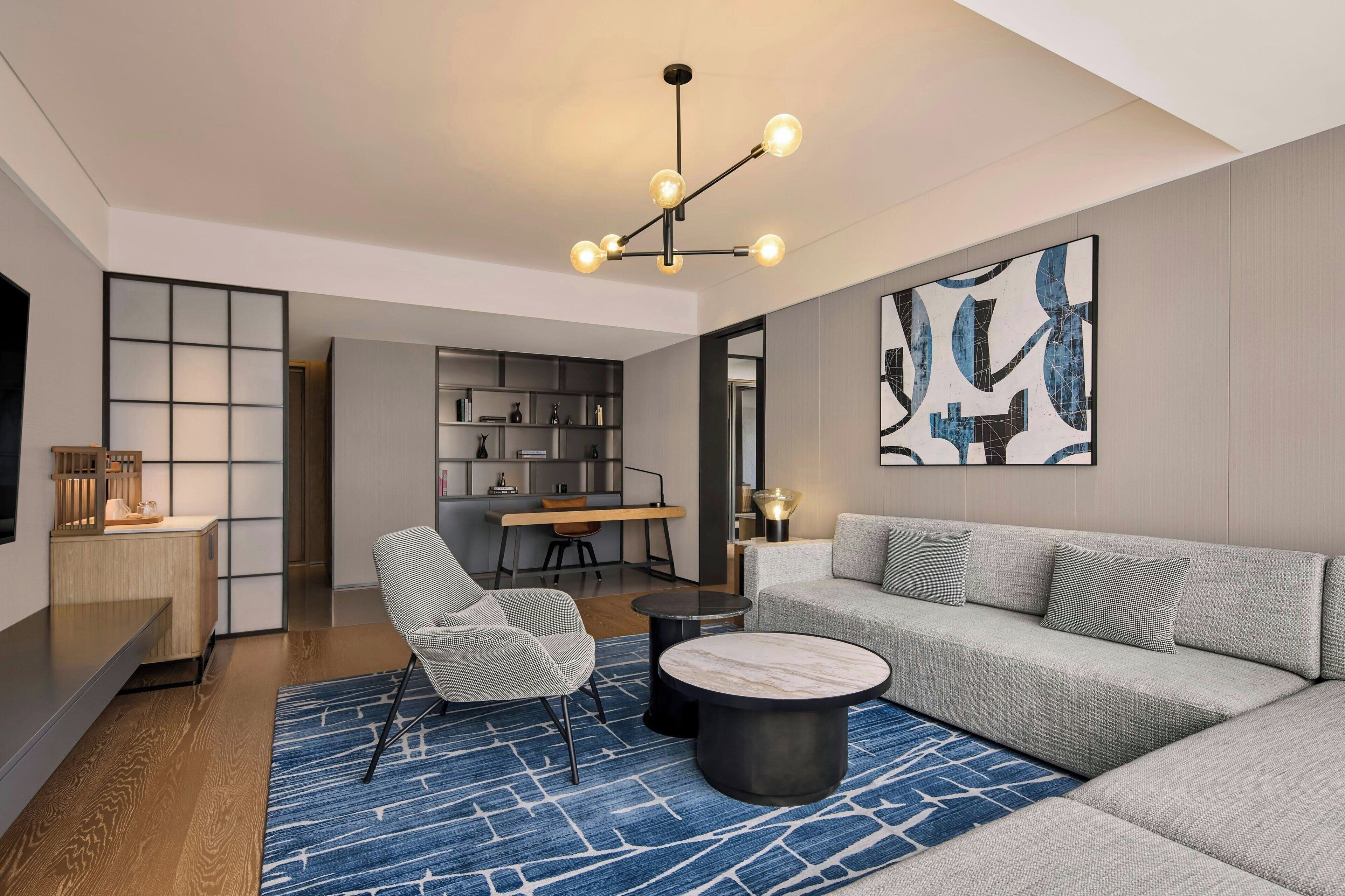 Een moderne woonkamer met een grijze bank, een ronde salontafel en een blauw gestreept vloerkleed. Er is een eethoek met een houten tafel en stoelen, en een keuken op de achtergrond.