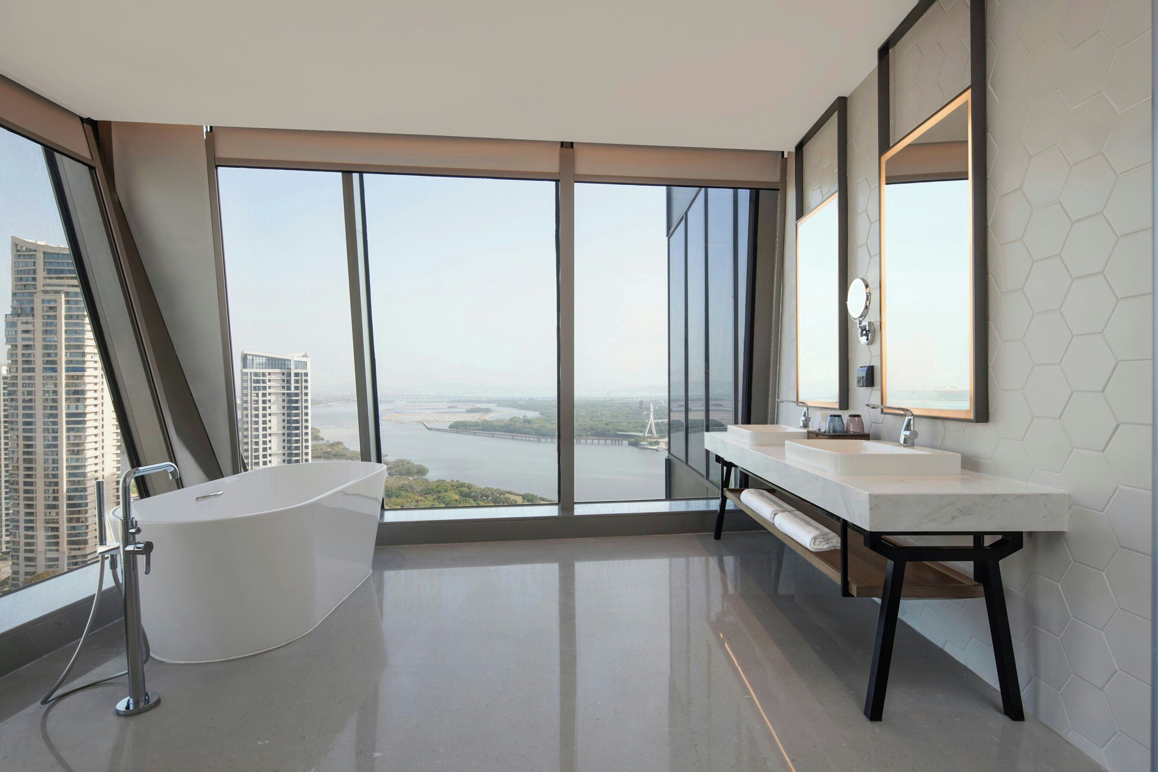 Een moderne badkamer met een groot bad, twee wastafels en een groot raam met uitzicht op de stad.