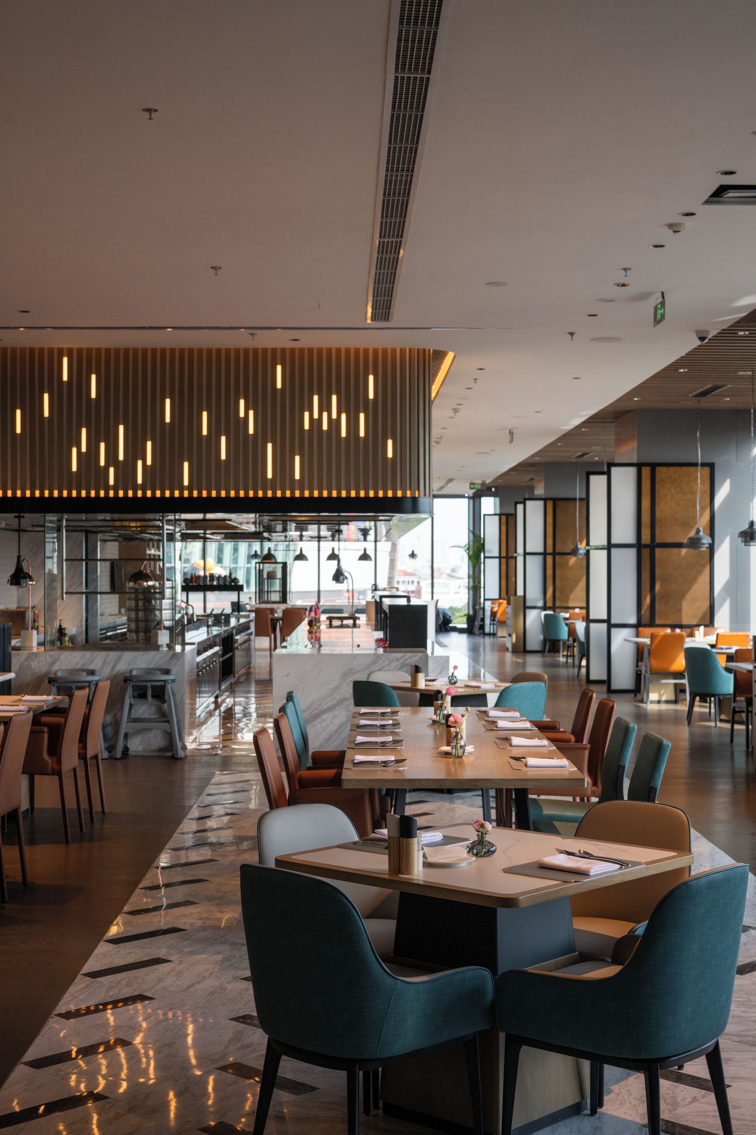 A modern restaurant with a long communal table, individual seating, and a kitchen area in the background.