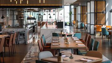 A modern restaurant with a long communal table, individual seating, and a kitchen area in the background.
