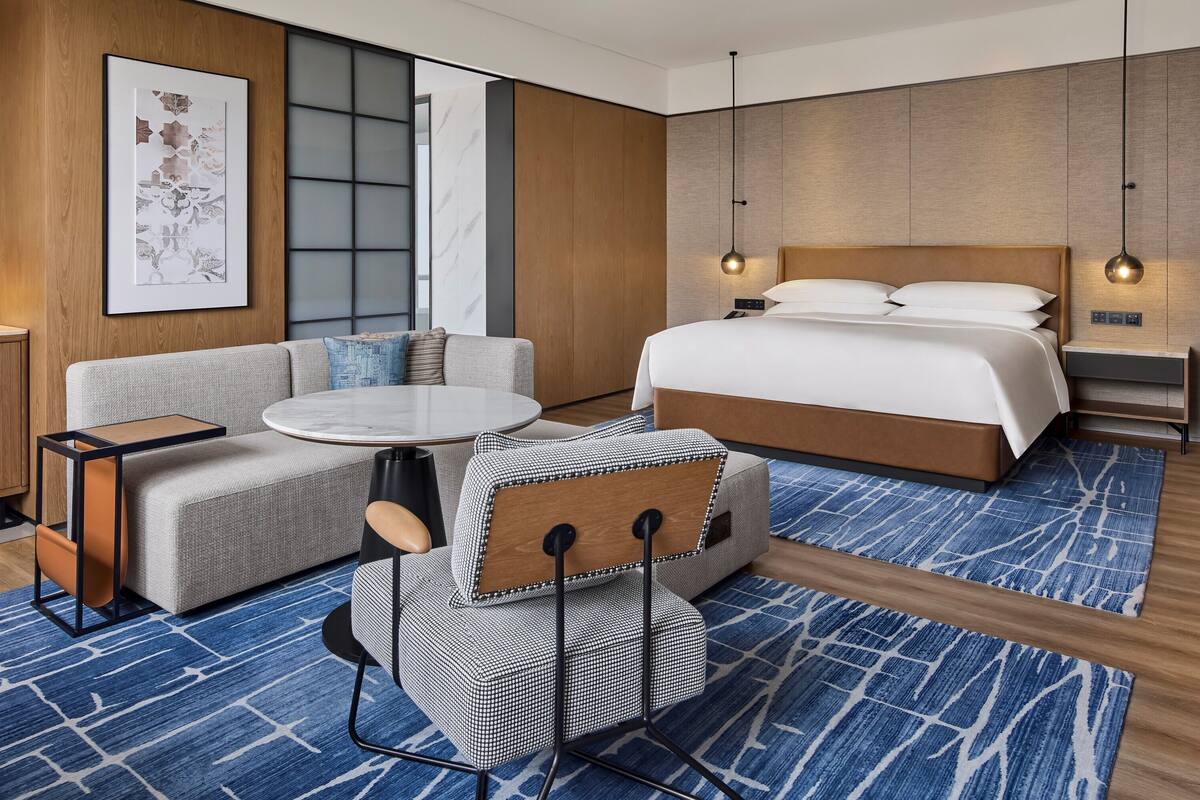 Ein Hotelzimmer mit einem Bett, einem Sofa, einem kleinen Tisch, einem Sessel, einem Schreibtisch mit Computer und einem großen Fenster.
