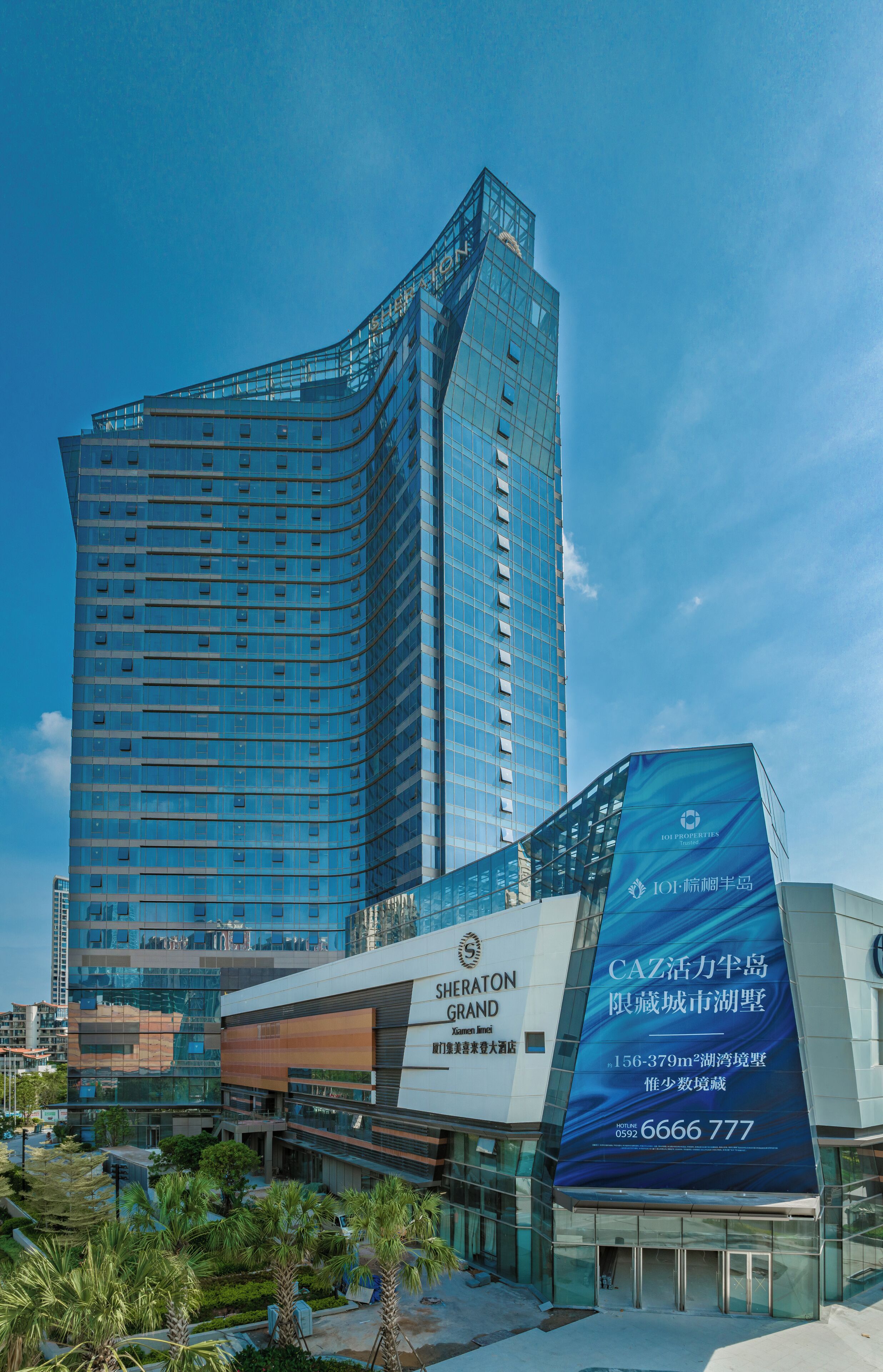 Een modern hotel met een opvallende glazen gevel, een Shersaton Grand bord en een grote advertentie voor CAZ.