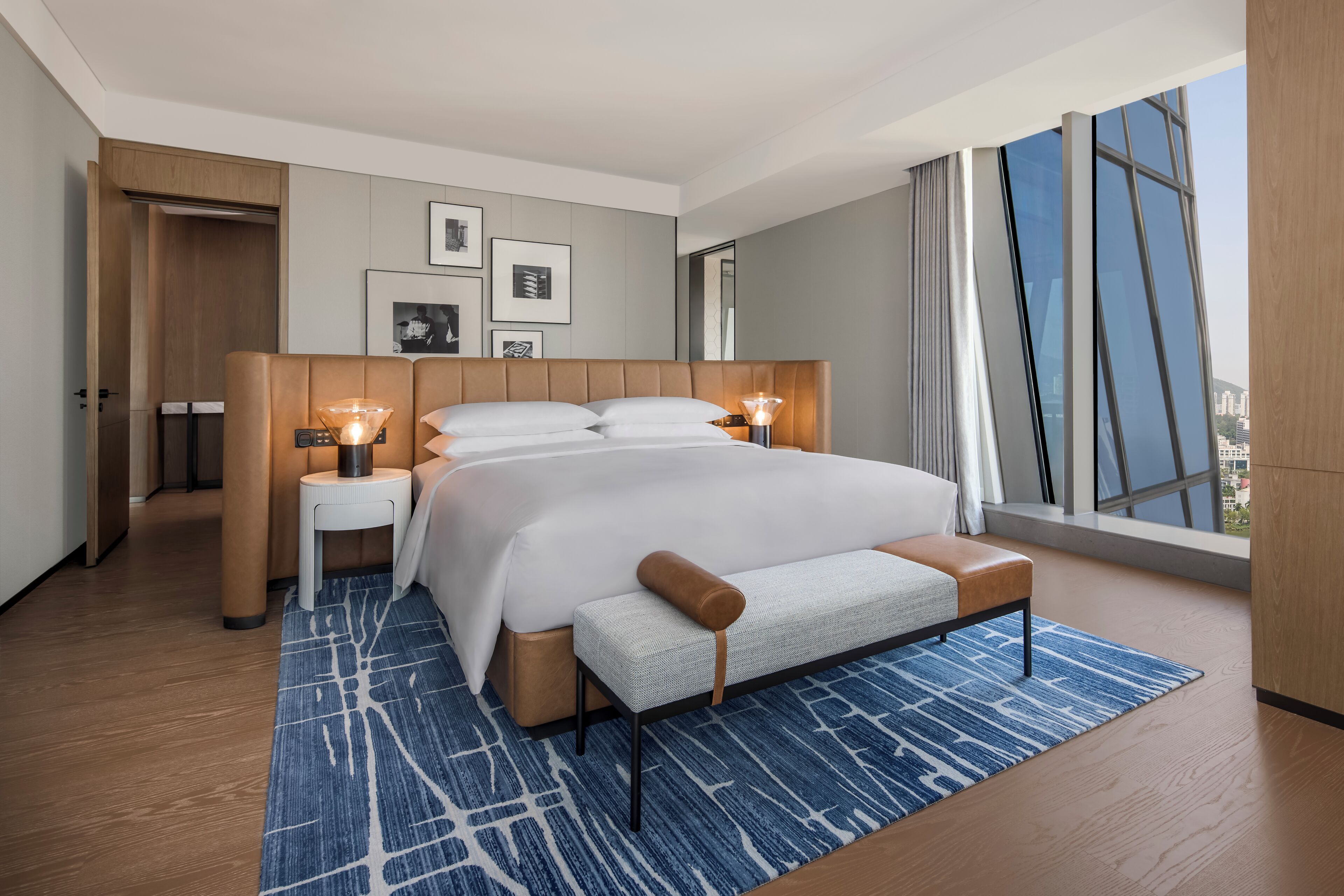Un dormitorio moderno con una cama grande, mesitas de noche con lámparas, un banco y vistas a la ciudad a través de amplios ventanales.
