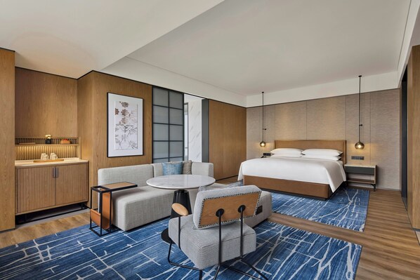 A modern hotel room with a bed, a sofa, a small table, and a chair. There is a large window and a wall-mounted artwork. - Sheraton Grand Xiamen Jimei (Xiamen)