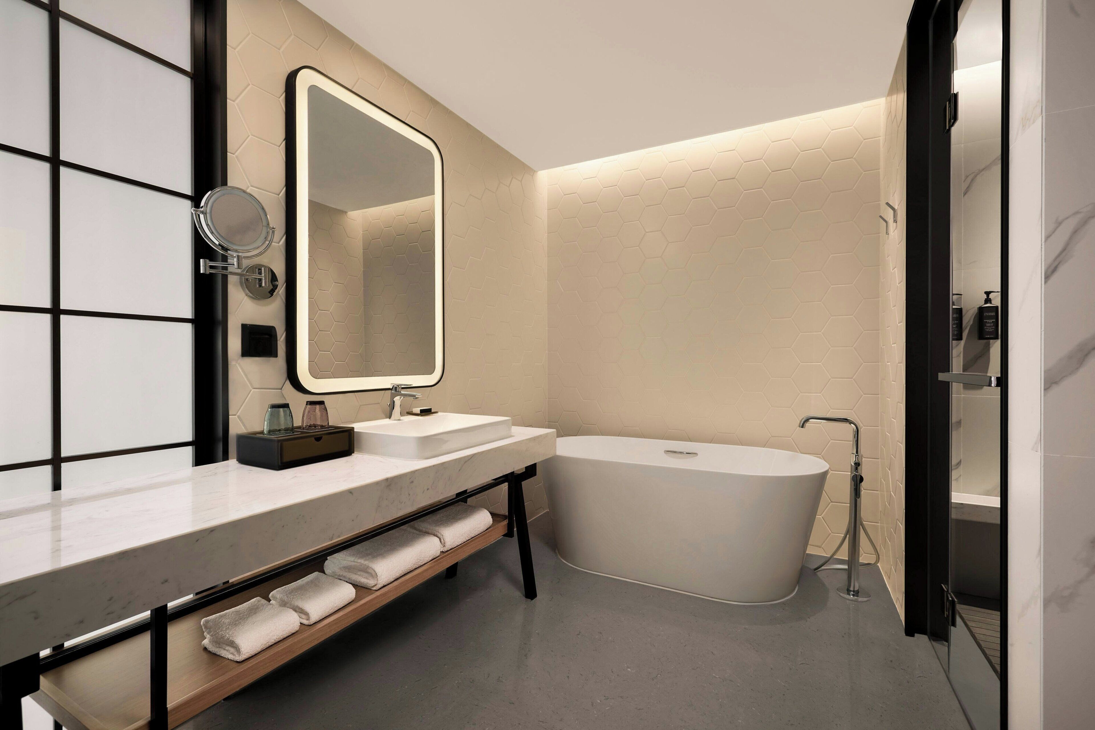 A modern bathroom with a large mirror, a bathtub, and a vanity with a shelf for towels.
