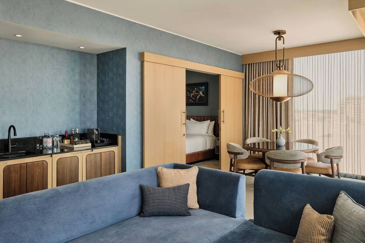 A modern hotel room with a blue sofa, a dining area, a kitchen, and a large window with curtains.