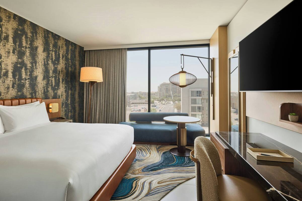 A hotel room with a large bed, a sofa, a small table, a TV, and a view of the city.