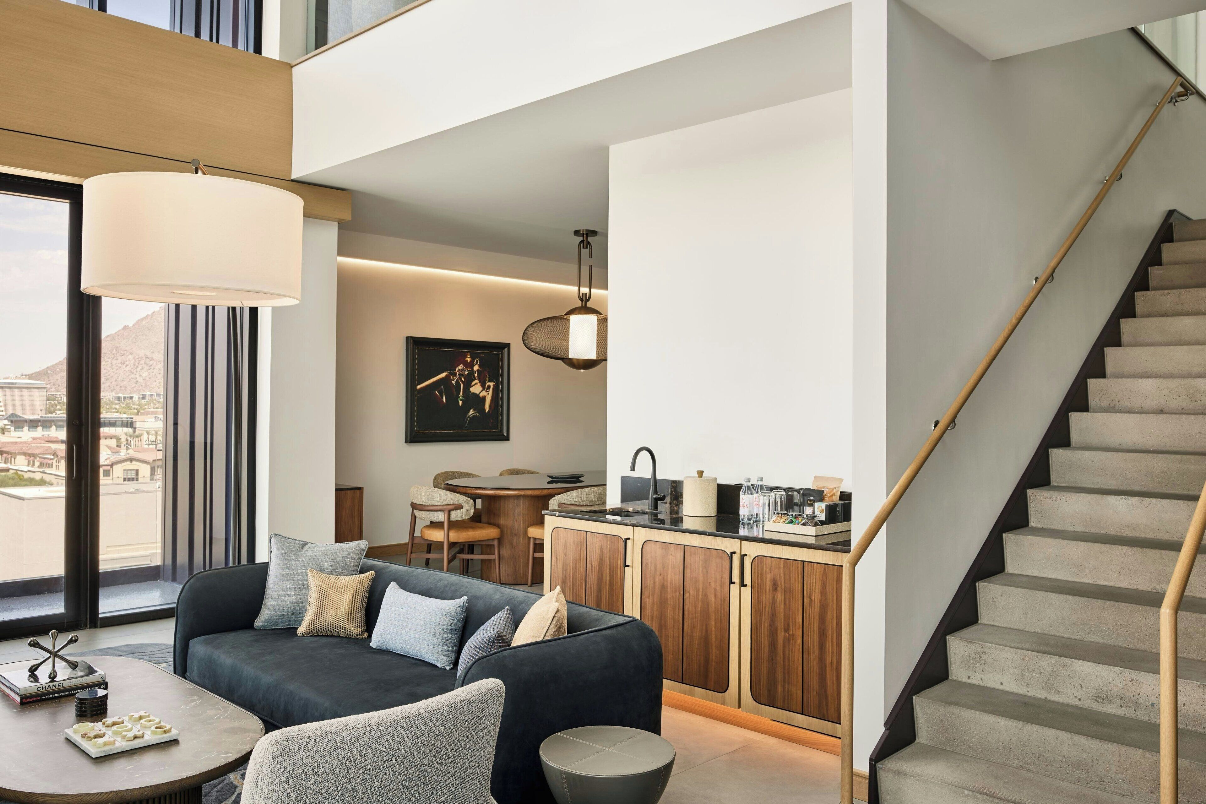 A modern living room with a sofa, coffee table, and a dining area. A kitchen with wooden cabinets and a staircase is visible.