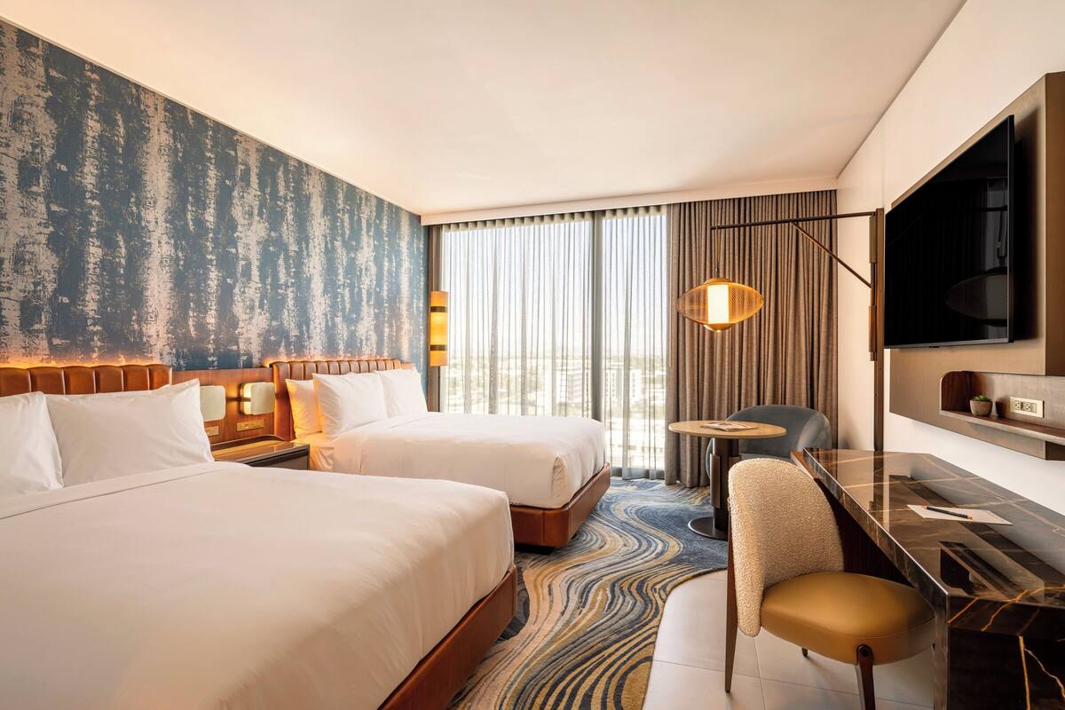 A hotel room with two beds, a desk, a chair, a TV, and a large window with curtains.