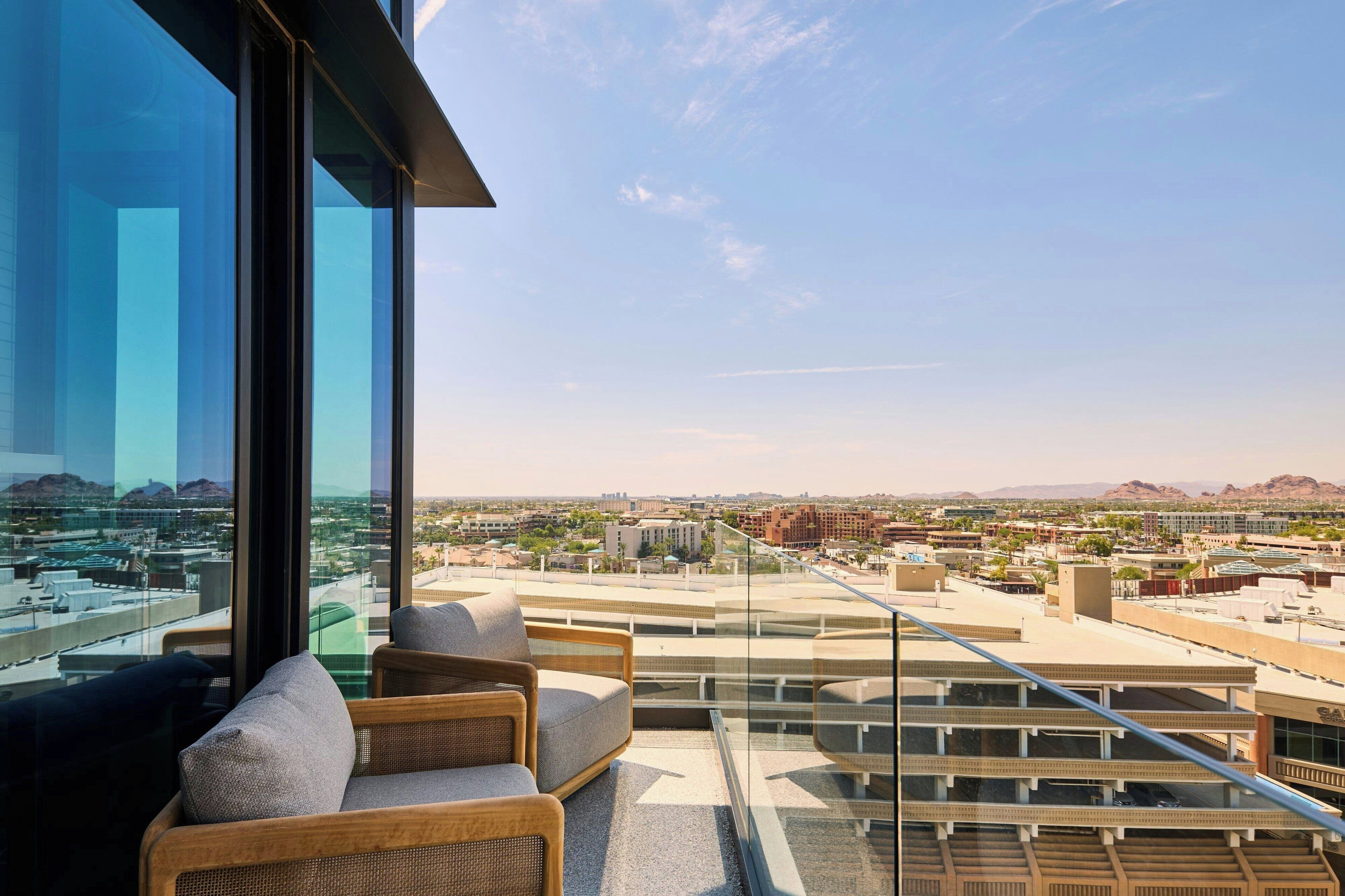 A balcony with wicker furniture and a glass railing overlooking a cityscape with buildings and mountains.