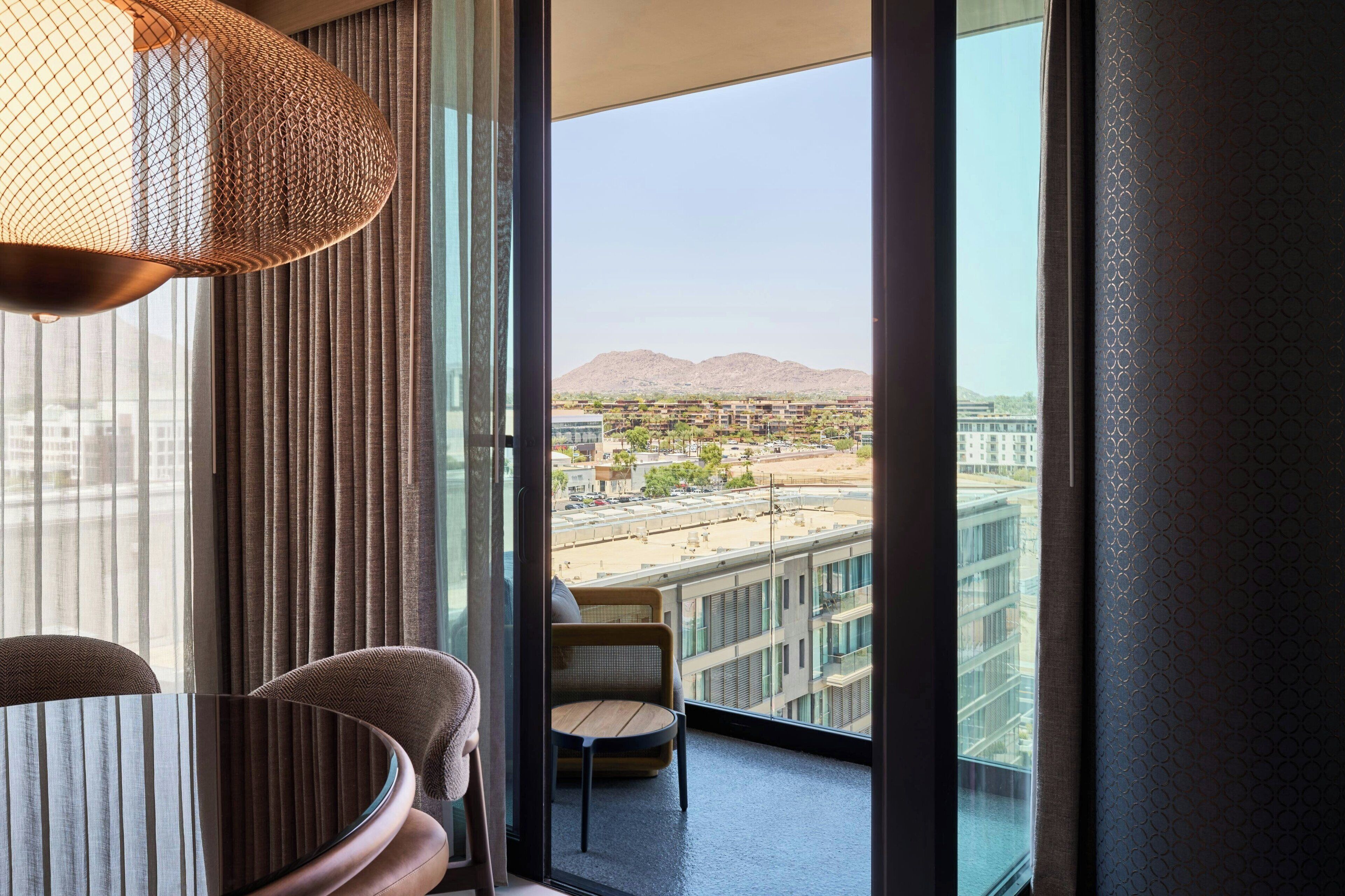 Habitación de hotel moderna con un ventanal amplio que ofrece vistas a la ciudad, una silla de mimbre y una mesa redonda.