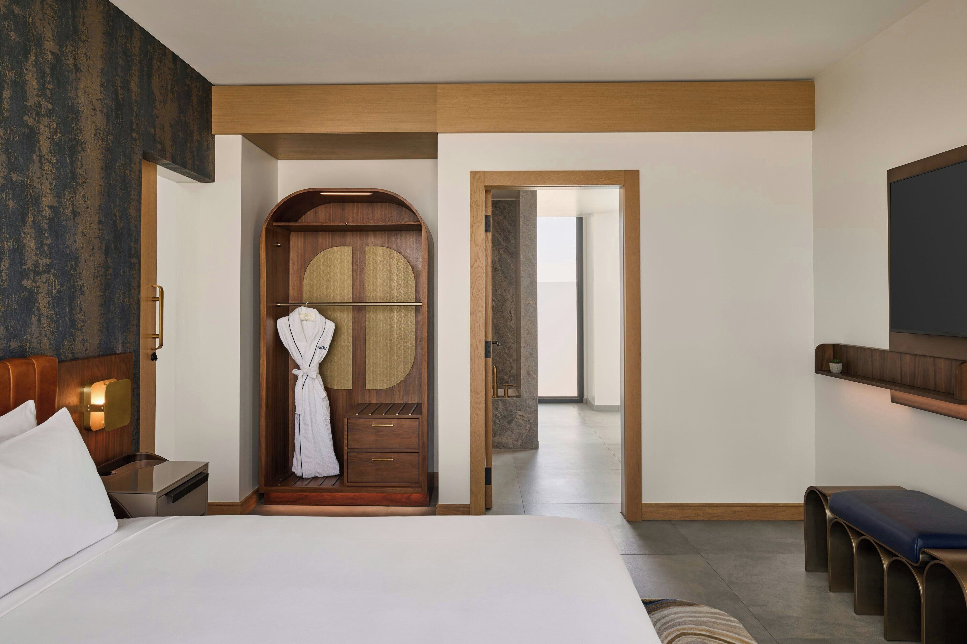 A hotel room with a large bed, a wooden wardrobe, a robe hanging on the door, and a bench by the window.