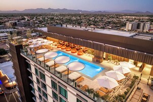 Uma área de piscina no terraço com várias espreguiçadeiras e guarda-sóis, cercada por um edifício moderno com telhado plano e vista para um horizonte urbano e montanhas ao fundo.