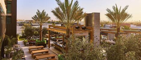 A rooftop bar with wooden tables and benches, surrounded by palm trees and potted plants.