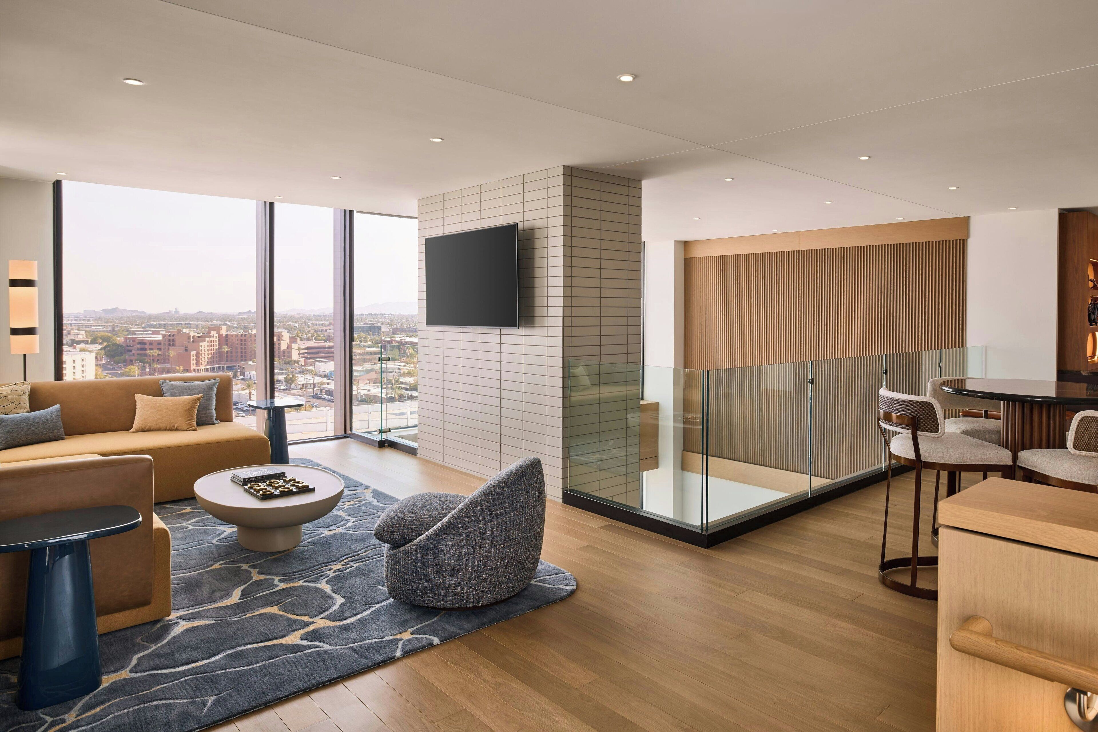 A modern hotel room with a large window, a flat-screen TV, a sofa, a coffee table, and a dining area with chairs.