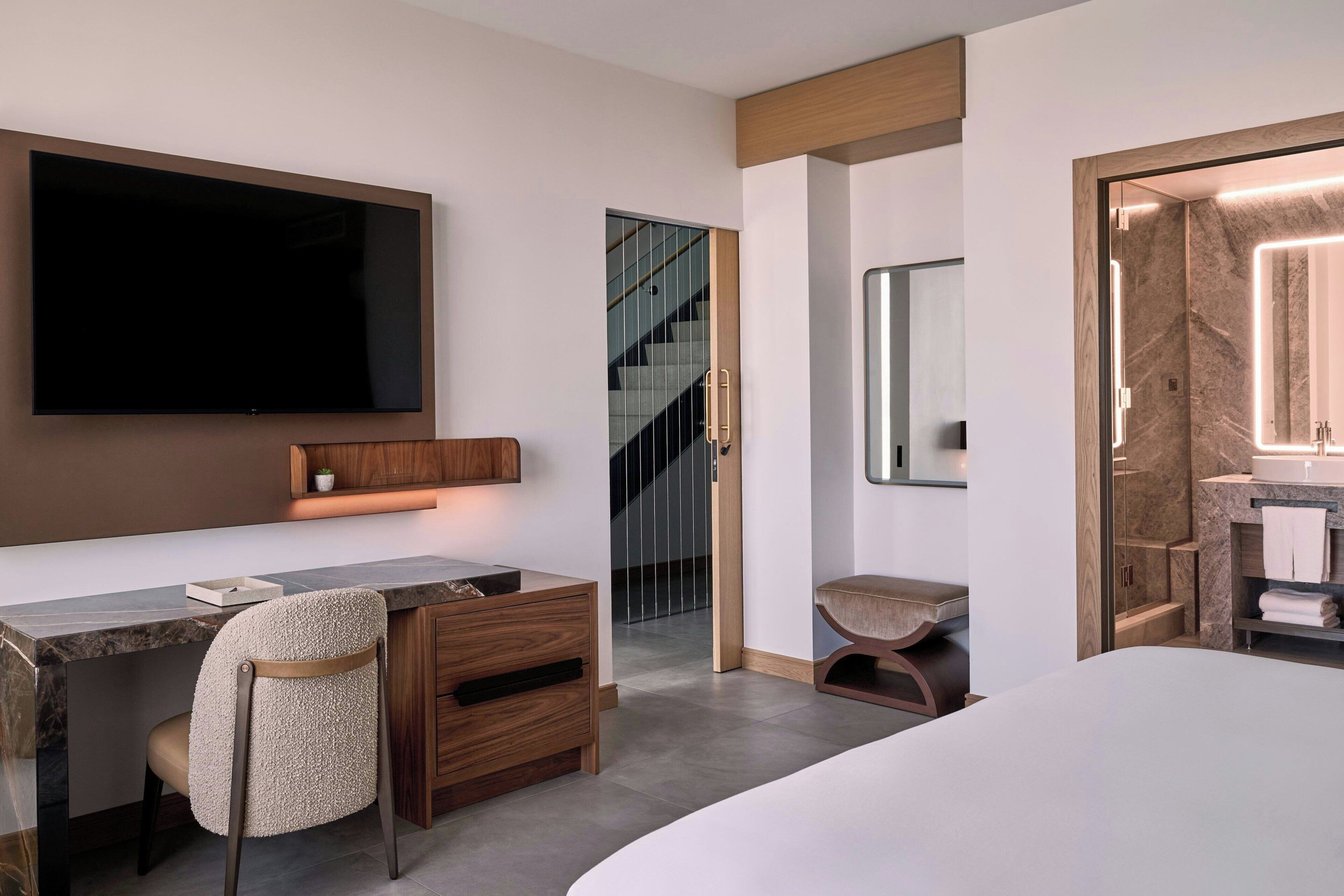 Ein modernes Hotelzimmer mit einem großen Bett, einem Schreibtisch mit Stuhl, einem an der Wand montierten Fernseher, einem Badezimmer mit Spiegel und Waschbecken sowie einer Treppe, die in eine andere Etage führt.