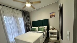 Habitación de hotel con cama, mesita de noche, lámpara, ventilador de techo y ventana con cortinas.