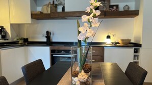 Et spisebord med en klar glasvase med hvide blomster og dekorative kugler, placeret i et moderne køkken med hvide skabe og træhylder.