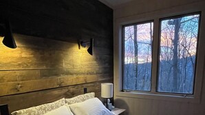 Een slaapkamer met een houten hoofdbord, een groot bed met beddengoed met een patroon, een nachtkastje met een lamp en een raam met uitzicht op bomen.