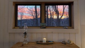 Een kamer met een houten tafel, een kaars en een raam met uitzicht op bomen en een zonsondergang.