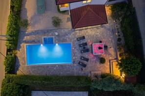 A rectangular swimming pool with a tiled deck, surrounded by a low hedge, and a patio area with outdoor furniture.