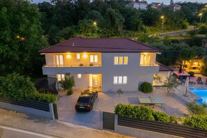 A two-story house with a car parked in the driveway, a swimming pool, and a well-lit outdoor area.