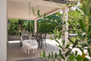 Een overdekt terras met een hangstoel en een tafel met stoelen.