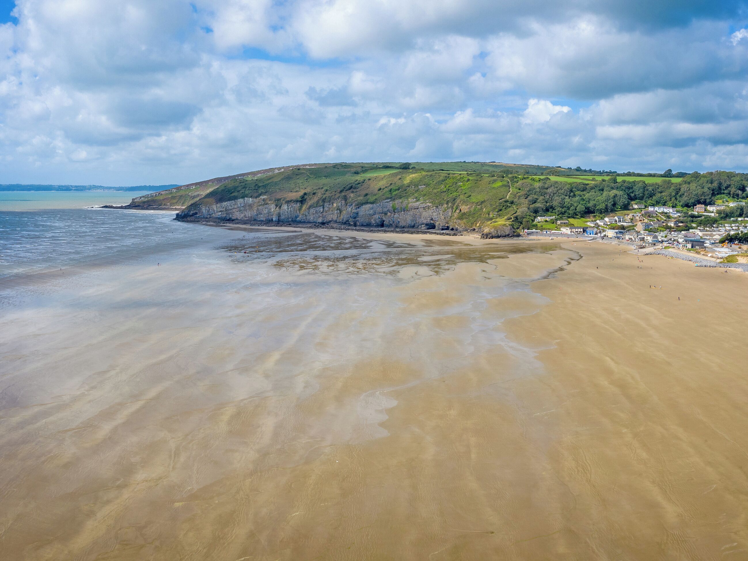 A beach with a sandy shore, a cliff with greenery, and a small settlement in the background.
