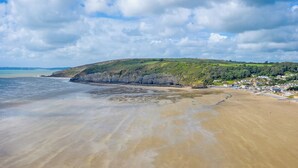A beach with a sandy shore, a cliff with greenery, and a small settlement in the background. - 4 bedroom accommodation in Carmarthen (Carmarthen)