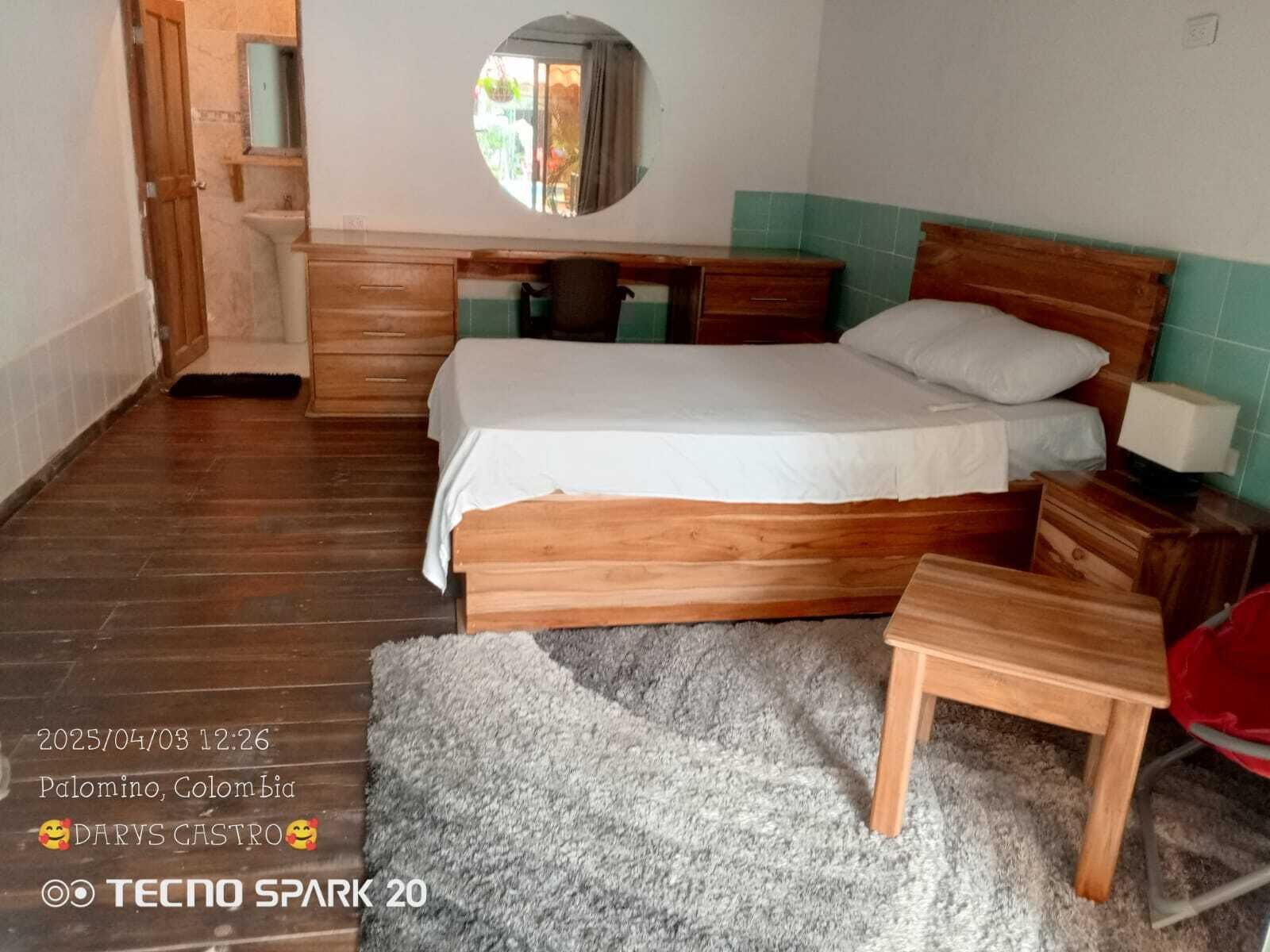 A bedroom with a wooden bed, a small bedside table, a mirror, and a window with a view of the outside.