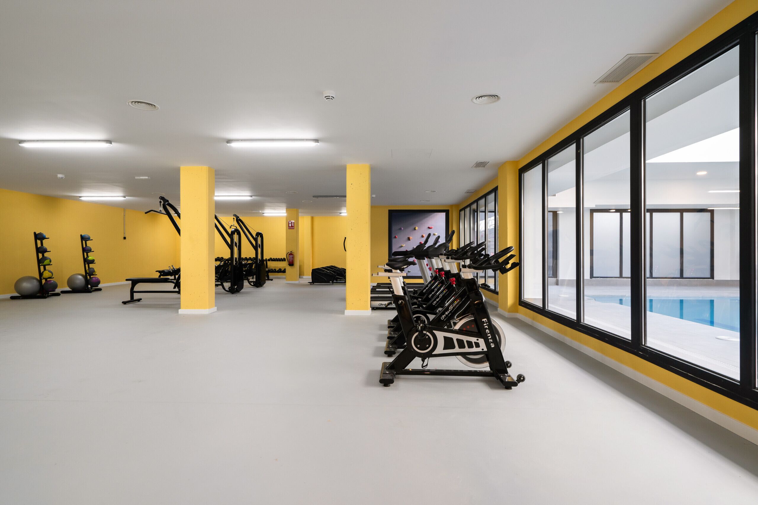 Une salle de sport équipée de matériel d’exercice, dotée de grandes fenêtres et arborant un décor aux couleurs jaune et blanc.