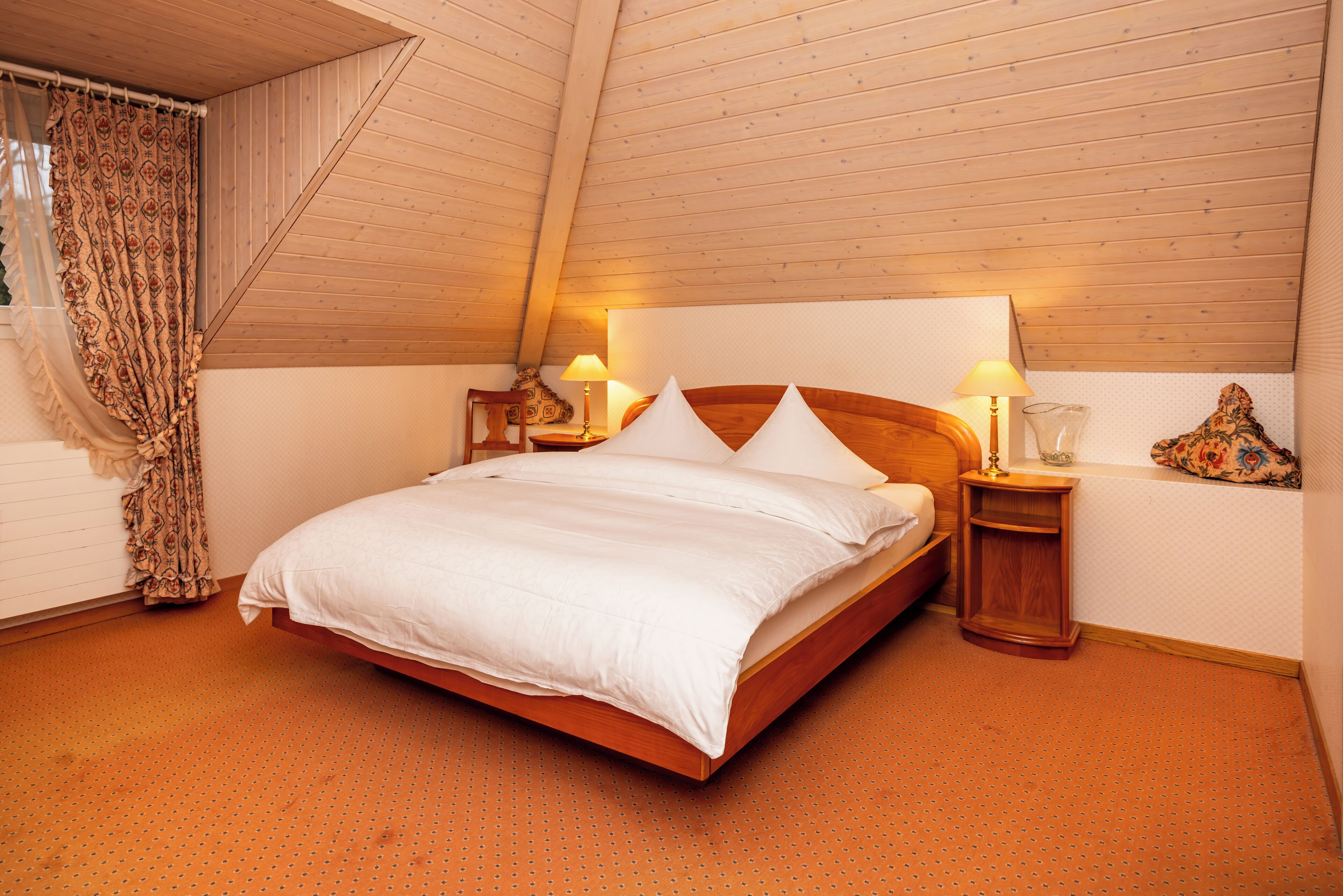 Ein Schlafzimmer mit Holzboden, einem Bett mit weißer Bettwäsche, zwei Nachttischen mit Lampen und einem gemusterten Vorhang.