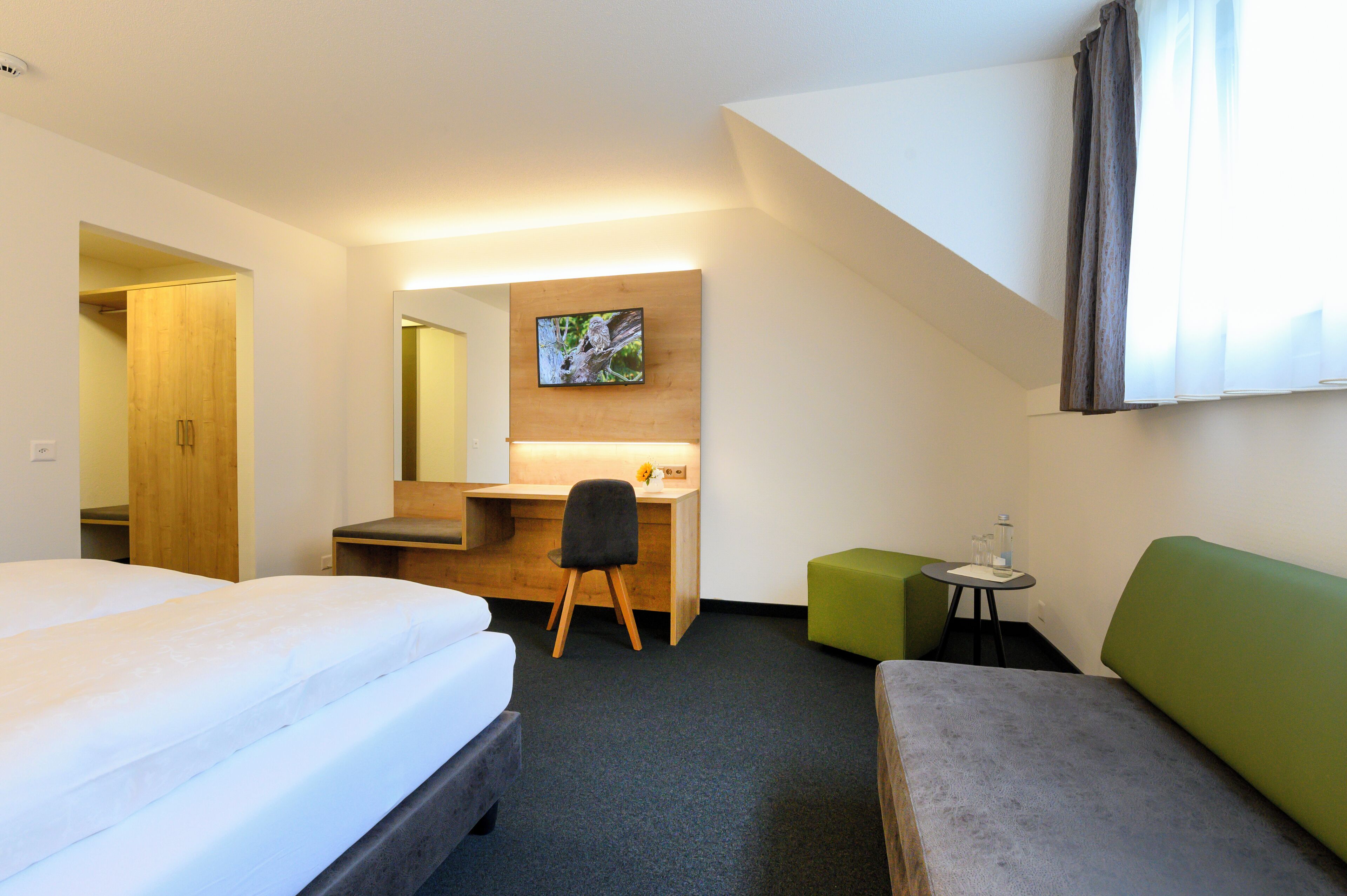 A hotel room with a bed, a desk with a chair, a small table with a glass, and a green ottoman.
