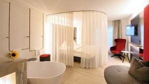 Una habitación de hotel moderna con una zona de ducha curvada, un sillón rojo, una mesita redonda y un televisor de pantalla plana montado en la pared.