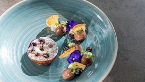 Un plato azul de cerámica con un postre cubierto de azúcar glas, arándanos y una flor morada.