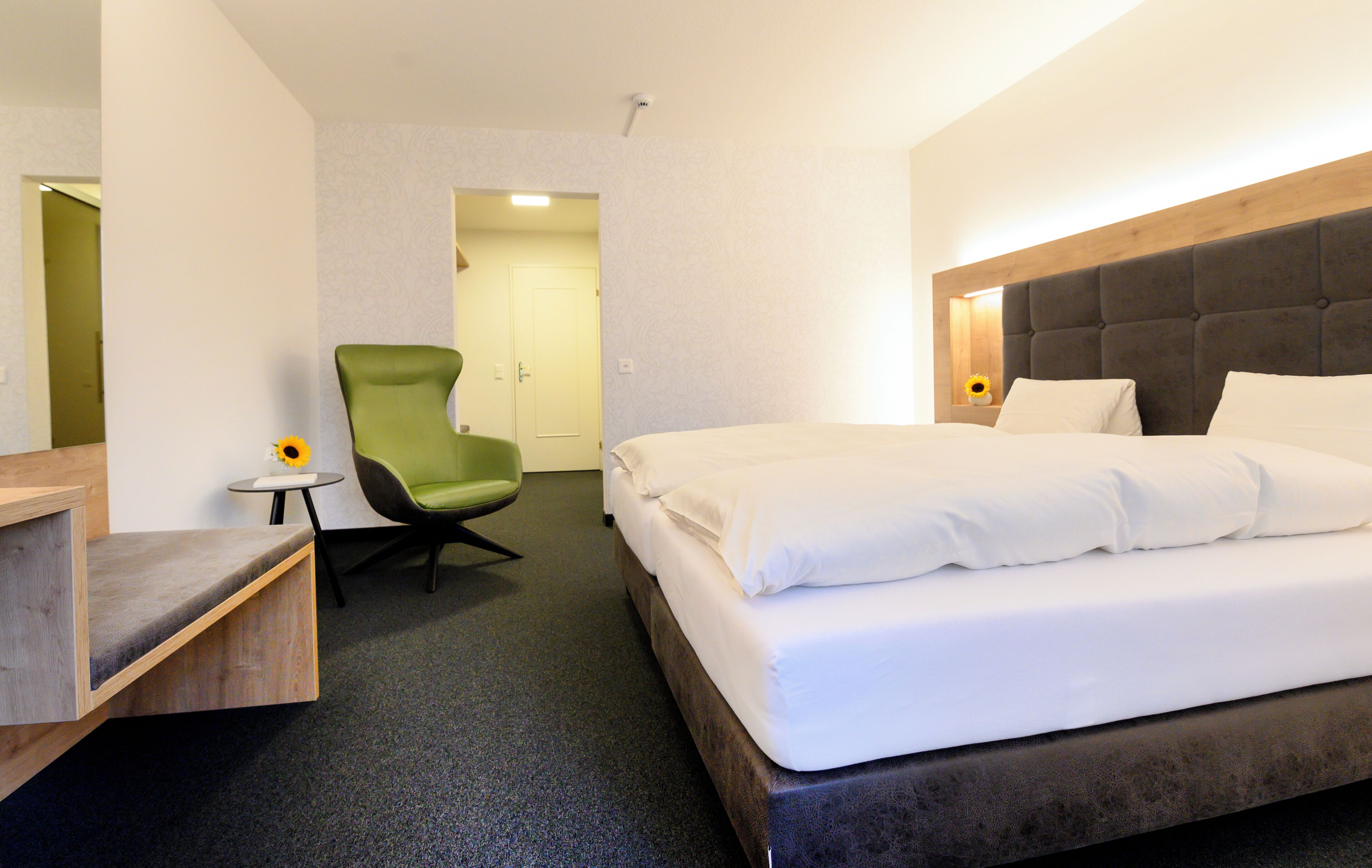 Ett hotellrum med en säng, en grön stol, ett litet bord och en dörr som leder till ett annat rum.