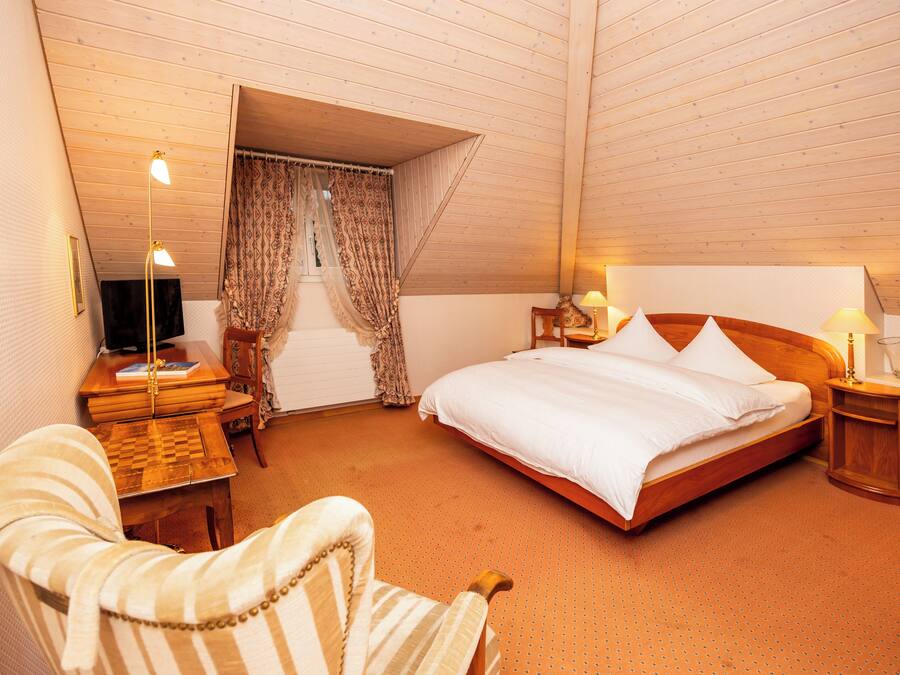 A bedroom with a wooden ceiling, a bed with white linens, a wooden nightstand with a lamp, a chair, and a small table with a book.