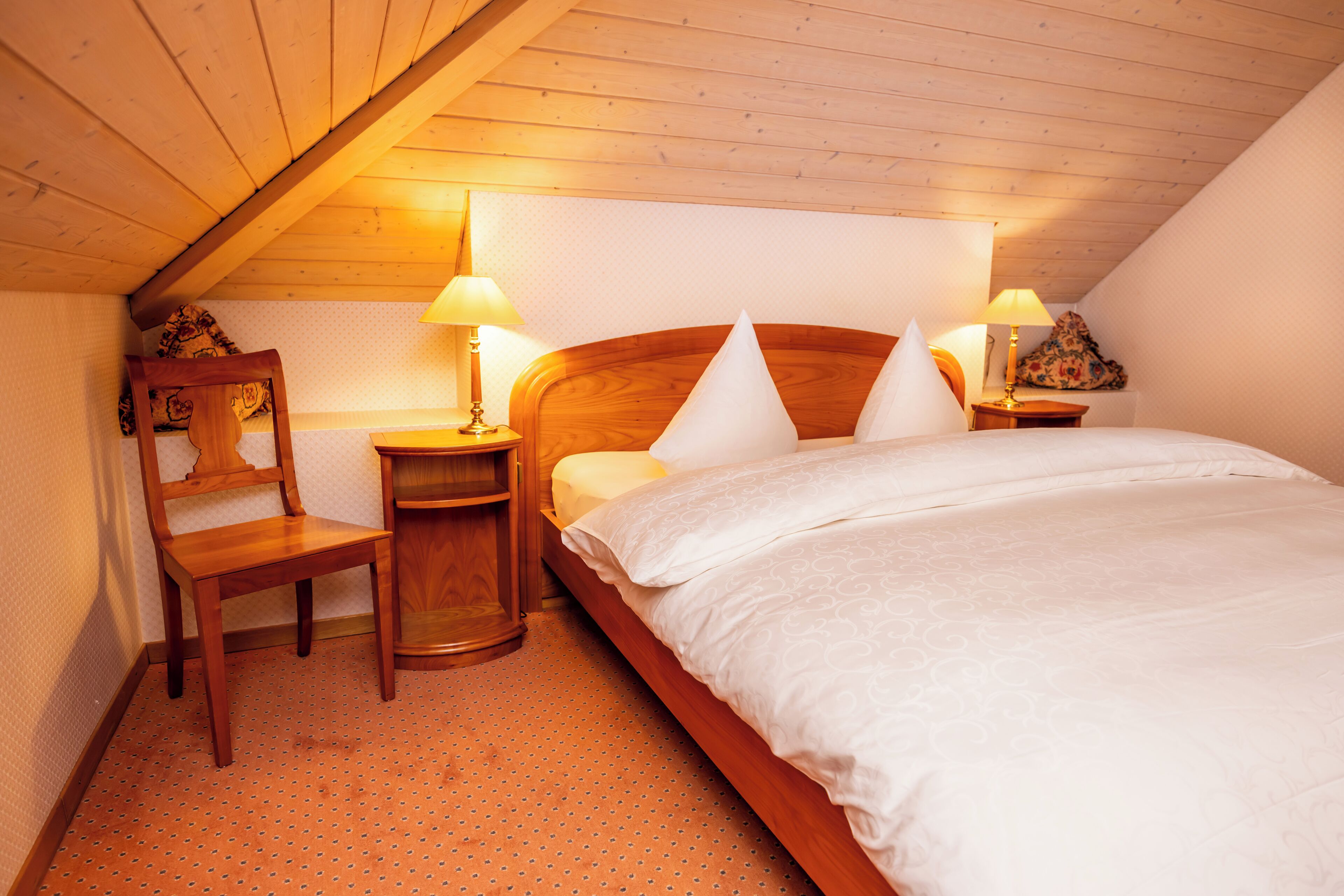 A bedroom with a wooden bed, two bedside tables with lamps, a wooden chair, and a patterned wall.