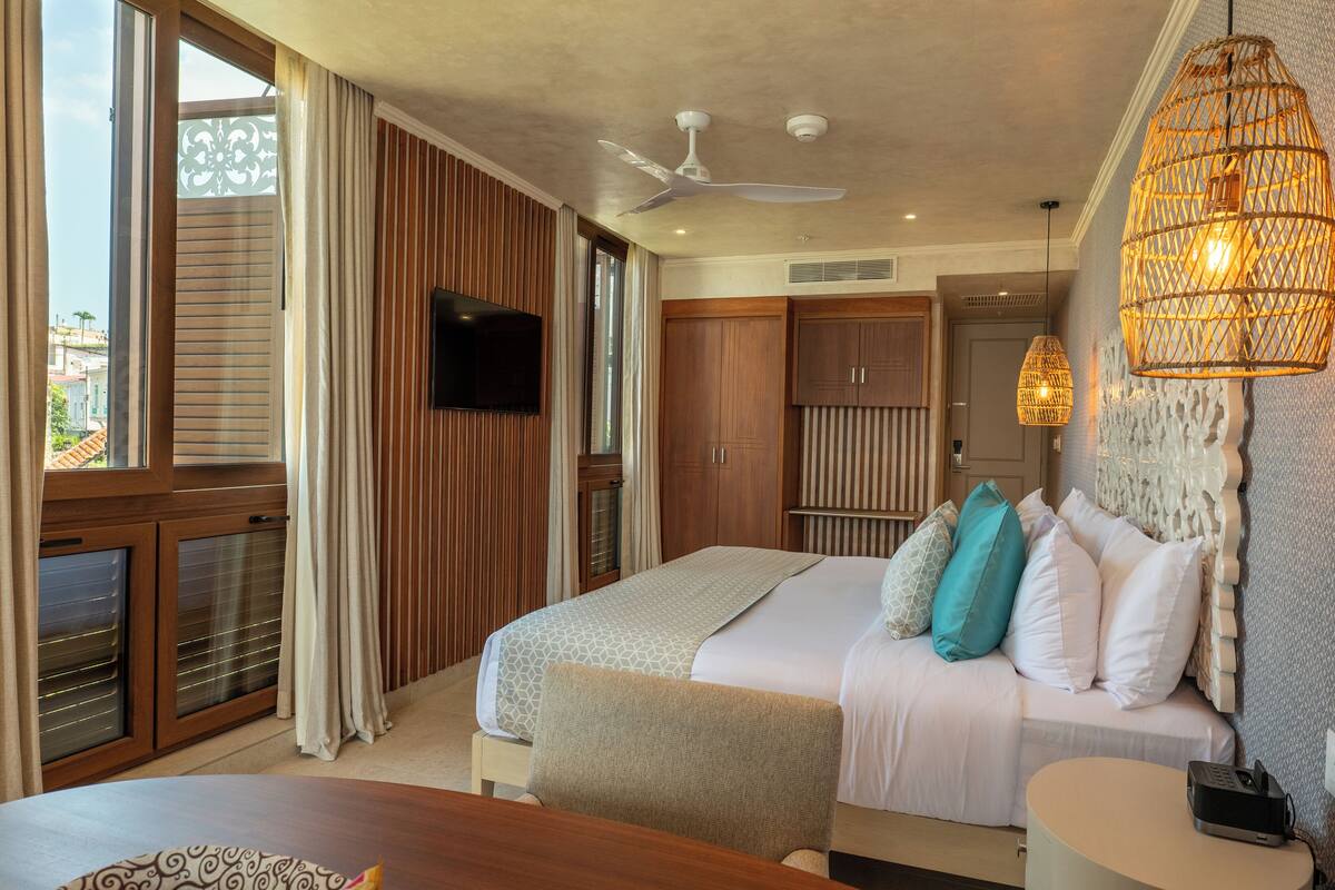 A hotel room with a bed, a TV, a ceiling fan, and a large window.