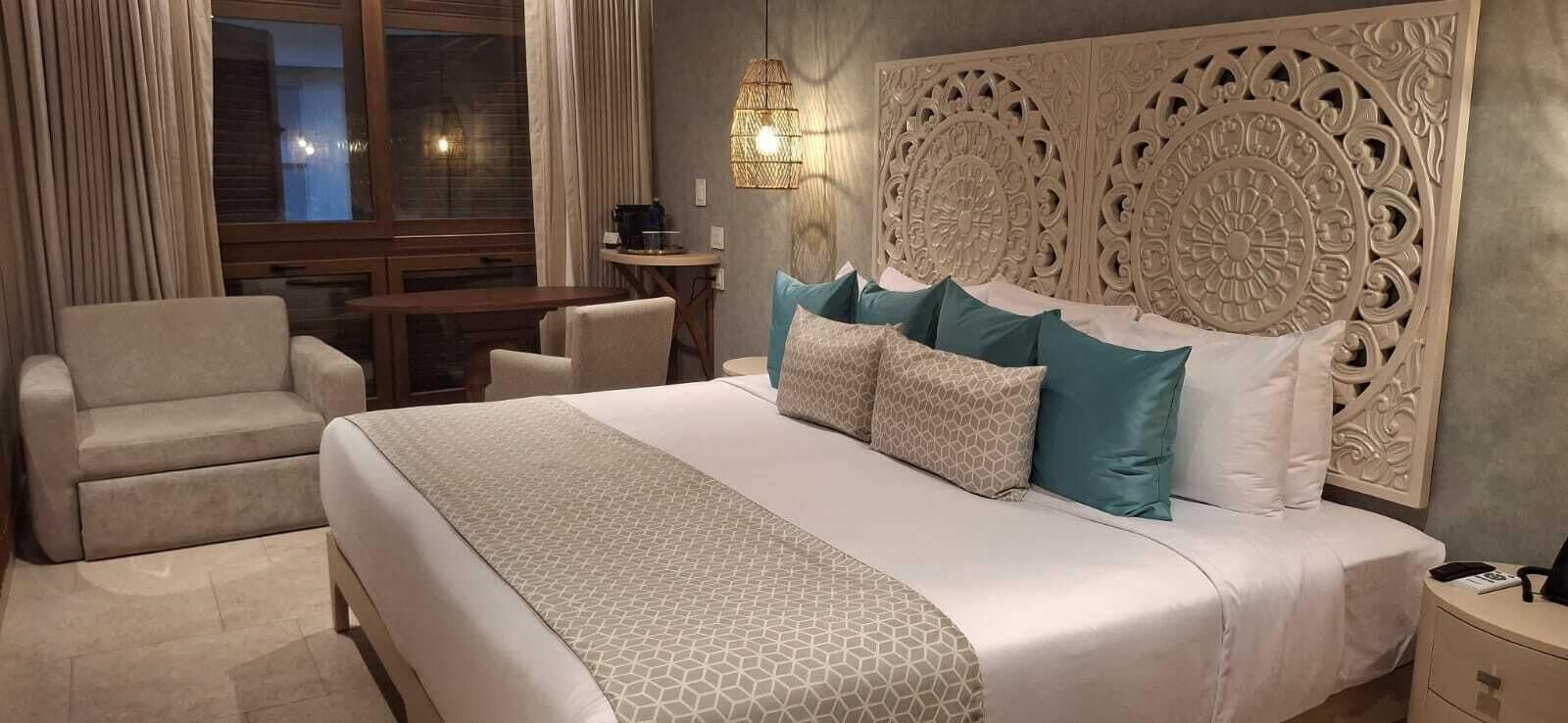 A hotel room with a large bed, a chair, a small table, and a decorative headboard.